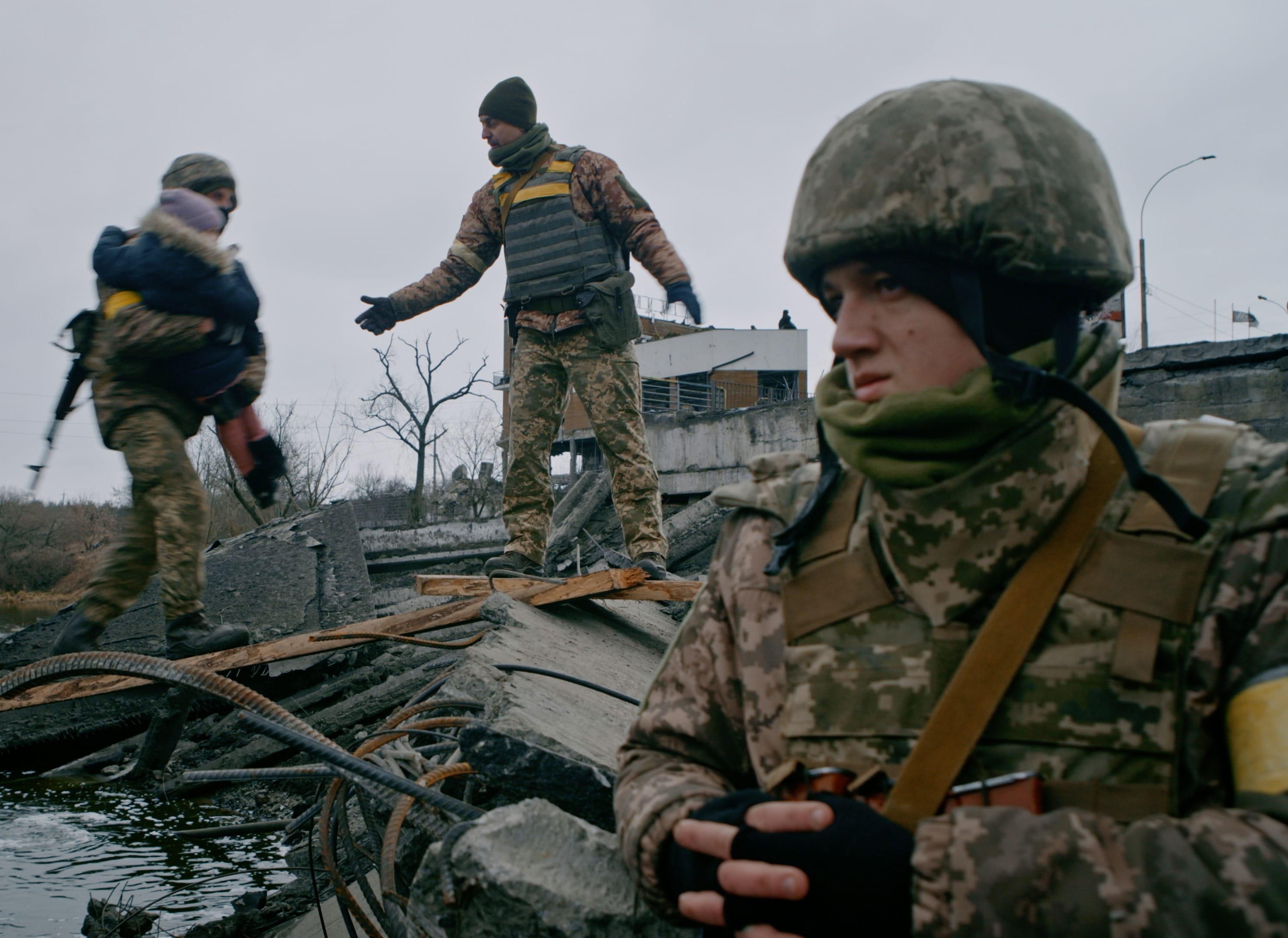 záběr z fronty s ozbrojeným vojákem v popředí a dvěma vojáky přenášejícími děti v pozadí